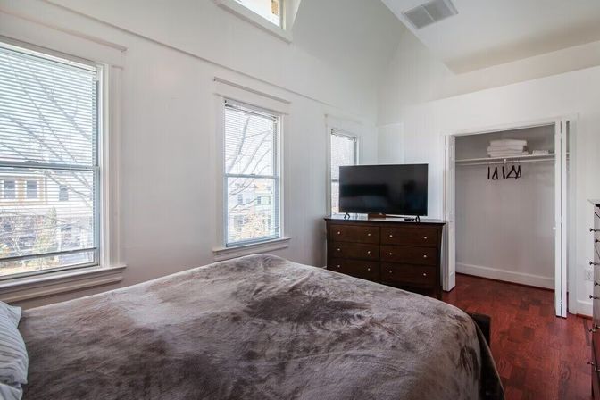 Sunny bedroom with three tall windows and blinds, plush gray bedspread, dark wood dresser with flat-screen TV, open closet with hangers and warm hardwood floors.