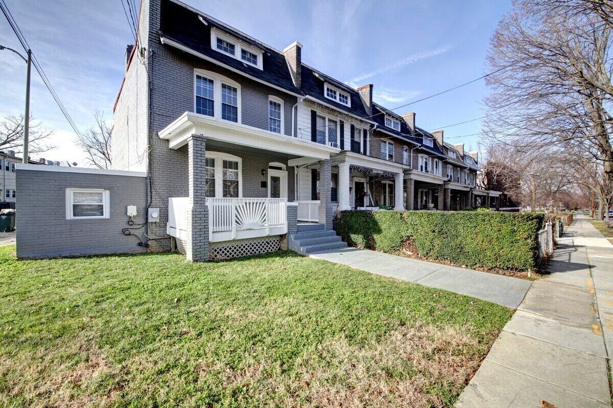Gray brick row houses with covered front porches, dormer windows and trimmed hedges along a tree-lined sidewalk in an urban residential neighborhood