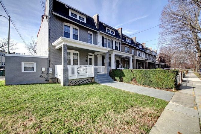 Gray brick row houses with covered front porches, dormer windows and trimmed hedges along a tree-lined sidewalk in an urban residential neighborhood