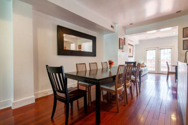 Sunlit open-plan dining room with a dark wooden table and eight mixed chairs on glossy hardwood floors, large rectangular wall mirror, small vase of flowers on the table, and French doors opening to a balcony.
