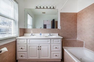 Bright residential bathroom with white double-sink vanity and storage cabinets, large mirror, beige tiled walls and built-in soaking tub next to a window with blinds
