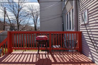 Red-painted backyard deck with a small red kettle grill and metal patio chair beside gray house siding, leafless trees and power lines in a suburban yard.