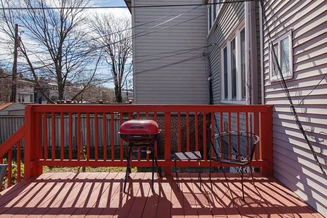 Red-painted backyard deck with a small red kettle grill and metal patio chair beside gray house siding, leafless trees and power lines in a suburban yard.