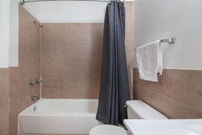 Cozy small bathroom with white bathtub, tan tile shower surround, dark gray textured shower curtain, chrome fixtures, white toilet and towel on wall-mounted rack