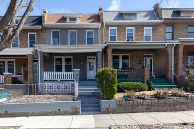 Two-story brick rowhouses with covered front porches, concrete steps, low retaining walls and small shrubbed front yards along a city sidewalk on a sunny day.