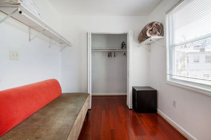 Sunlit compact apartment bedroom with hardwood floors, red-accent sofa, open closet, wall shelves and a window with blinds