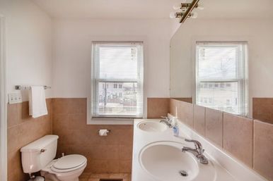 Sunlit residential bathroom with double vanity and chrome faucets, toilet, beige tile wainscoting, large mirror and two windows with blinds.