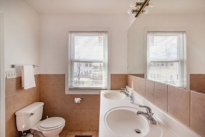 Sunlit residential bathroom with double vanity and chrome faucets, toilet, beige tile wainscoting, large mirror and two windows with blinds.