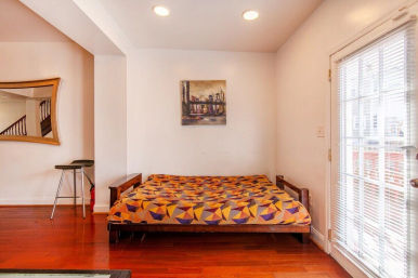 Bright urban bedroom nook with low wooden platform bed topped by orange and purple geometric quilt, polished hardwood floors, white walls with cityscape painting, curved wall mirror and stool to the left, glass door with blinds to the right.