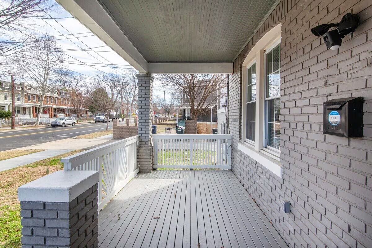 Cozy gray-brick front porch with white railing and painted wood floor, security camera box on the wall, overlooking a tree-lined residential street with rowhouses.