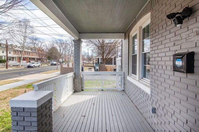 Cozy gray-brick front porch with white railing and painted wood floor, security camera box on the wall, overlooking a tree-lined residential street with rowhouses.