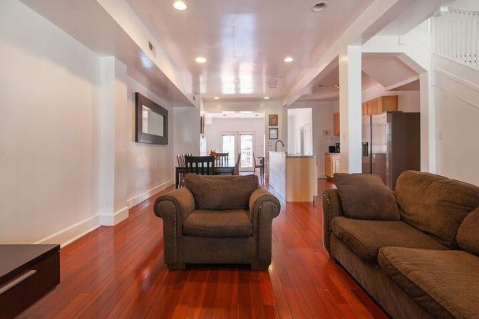 Cozy open-concept living room with cherry hardwood floors, brown upholstered sofa and armchair, dining table, recessed lighting and a modern kitchen with stainless steel refrigerator in the background.