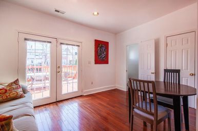 Bright living-dining room with French doors opening to a sunlit wooden deck, warm hardwood floors, round dining table, sofa with colorful pillows, white walls and red abstract art.