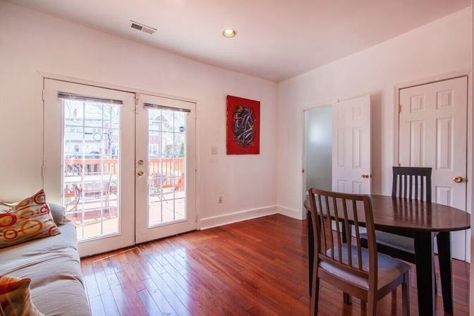 Bright living-dining room with French doors opening to a sunlit wooden deck, warm hardwood floors, round dining table, sofa with colorful pillows, white walls and red abstract art.