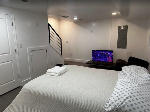 Finished basement bedroom with white walls and recessed lighting, modern black metal stair railing, neatly made bed with folded towels and flat-screen TV on a low stand.