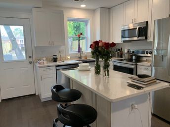 Bright modern white kitchen with marble island and two black bar stools, stainless steel appliances, sink under window, and vases of red roses on the counter.