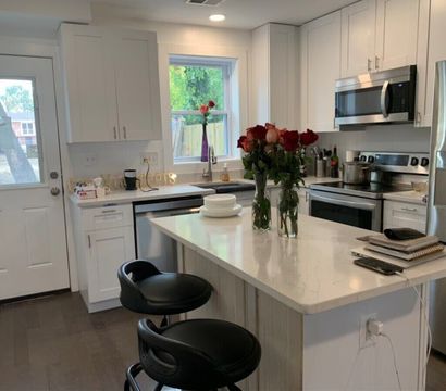 Bright modern white kitchen with marble island and two black bar stools, stainless steel appliances, sink under window, and vases of red roses on the counter.