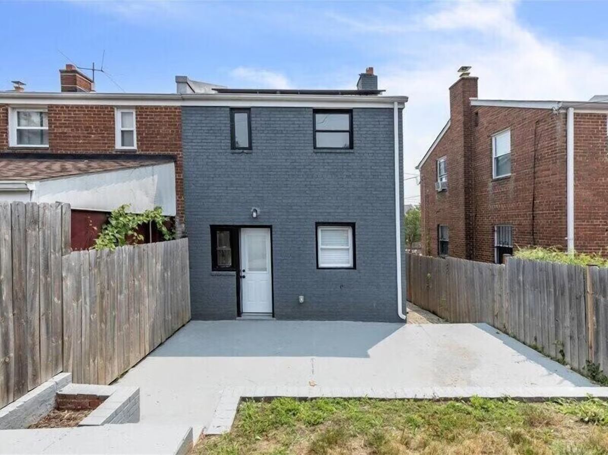 Urban two-story gray-painted brick rowhouse with a white back door and windows opening to a clean concrete backyard patio, flanked by wooden privacy fences and neighboring red-brick homes.