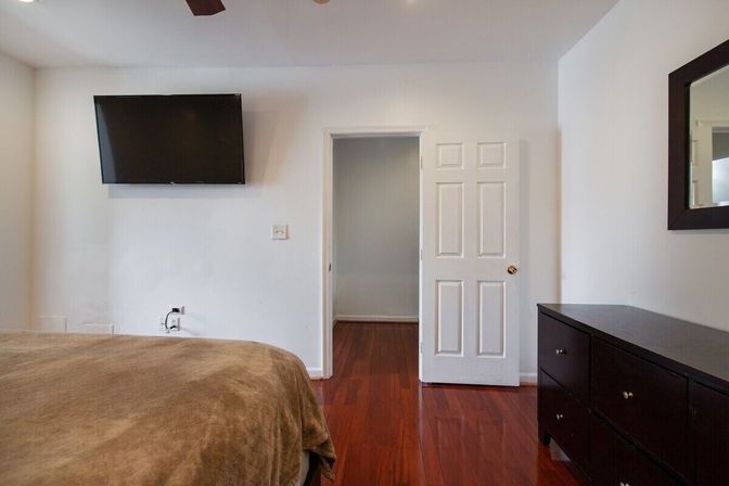 Cozy minimalist bedroom with brown bedspread, wall-mounted flat-screen TV, white walls, glossy hardwood floors, open white door and dark wood dresser with mirror.