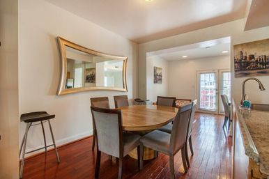 Sunlit open-plan dining room with round wooden table, six gray upholstered chairs, large curved mirror, hardwood floors and French doors to a balcony.