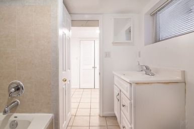 Bright compact home bathroom with beige-tiled bathtub and chrome fixtures, white vanity and mirror, narrow window with blinds, and open door leading to a tiled hallway with louvered closet doors.