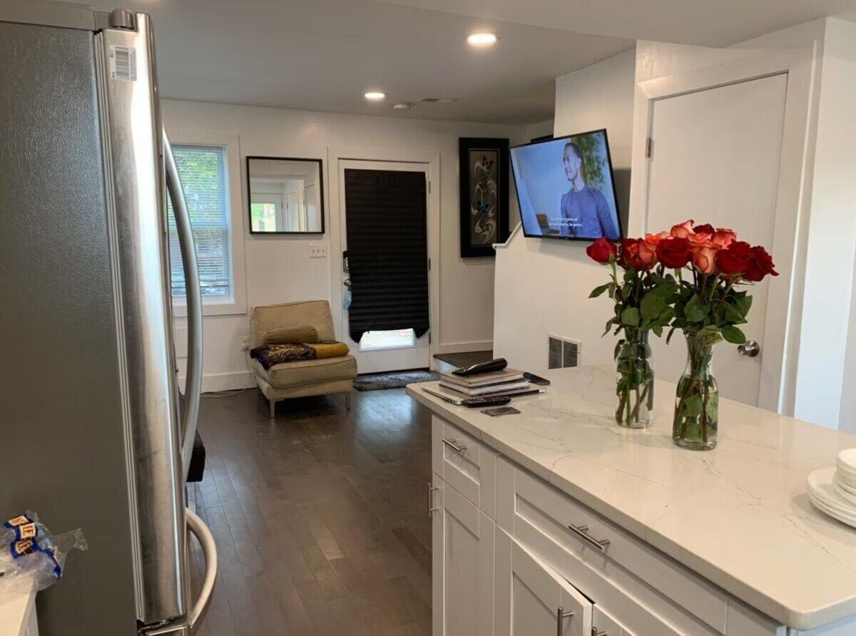Open-concept modern kitchen and living area with stainless steel refrigerator, white shaker cabinets and marble-look quartz island topped with two vases of red roses, dark hardwood floors, a cozy armchair by a back door with a black blind, and a wall-mounted TV showing a person.