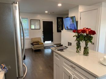 Open-concept modern kitchen and living area with stainless steel refrigerator, white shaker cabinets and marble-look quartz island topped with two vases of red roses, dark hardwood floors, a cozy armchair by a back door with a black blind, and a wall-mounted TV showing a person.