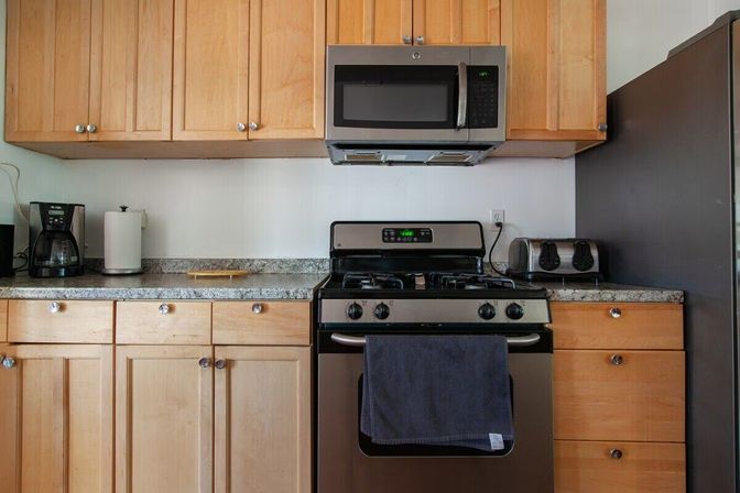 Cozy kitchen with light wood cabinets, stainless steel gas stove and over‑the‑range microwave, granite countertop, coffee maker, toaster and navy towel on the oven