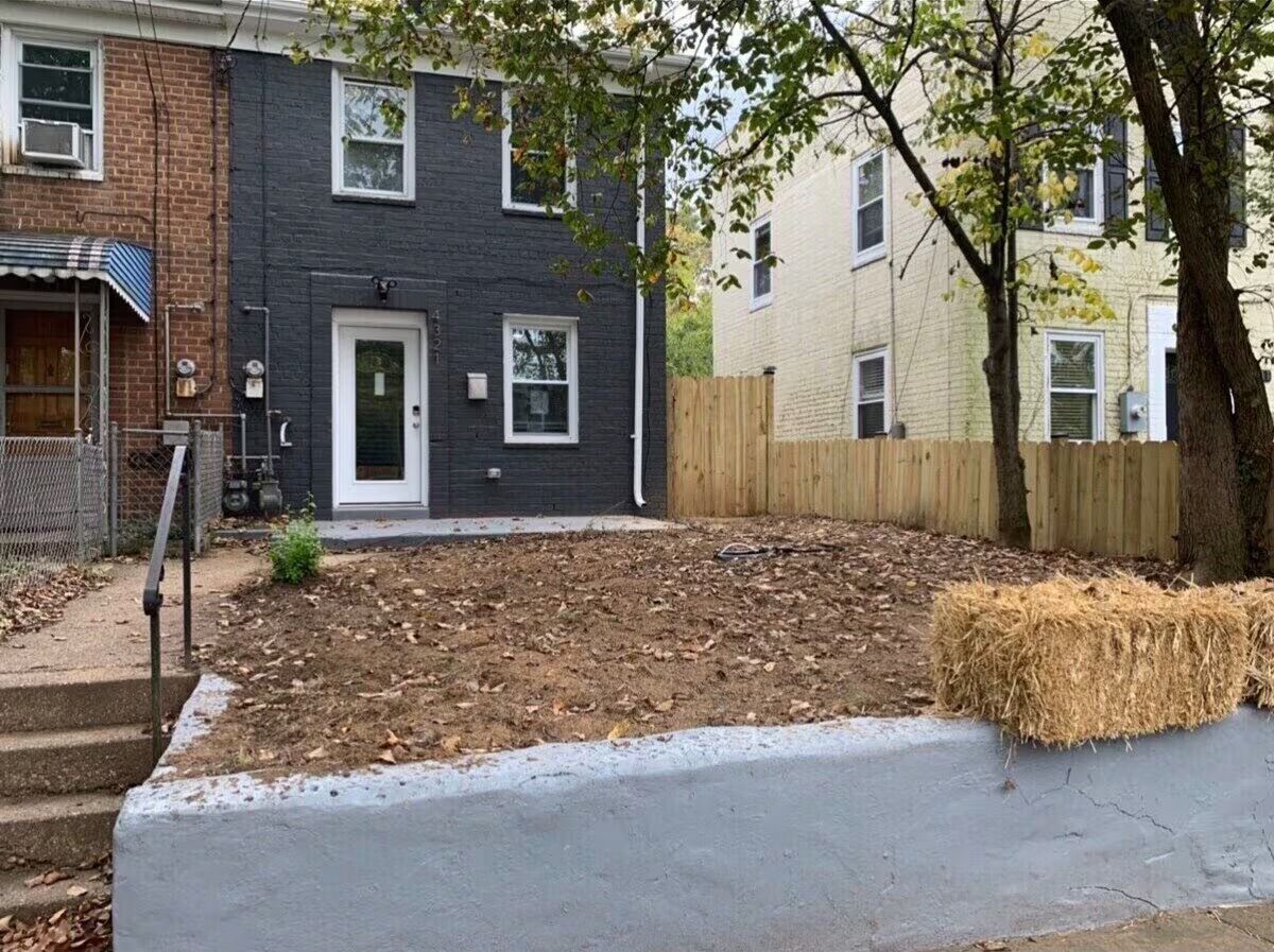 Urban brick rowhouse with dark gray painted facade and glass front door, small raised front yard of bare soil and fallen leaves behind a concrete retaining wall, sidewalk steps, wooden fence, trees and a hay bale