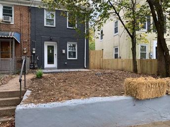 Urban brick rowhouse with dark gray painted facade and glass front door, small raised front yard of bare soil and fallen leaves behind a concrete retaining wall, sidewalk steps, wooden fence, trees and a hay bale