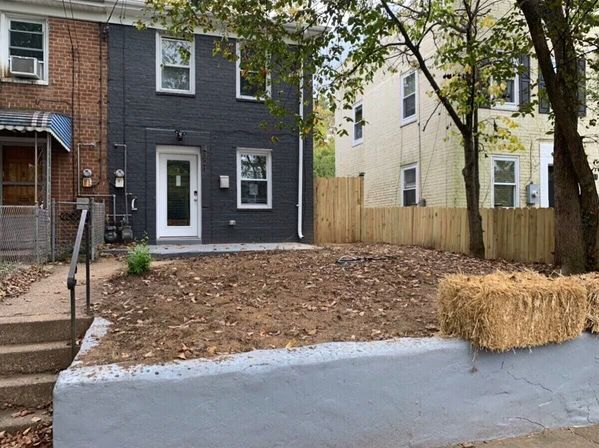 Urban brick rowhouse with dark gray painted facade and glass front door, small raised front yard of bare soil and fallen leaves behind a concrete retaining wall, sidewalk steps, wooden fence, trees and a hay bale