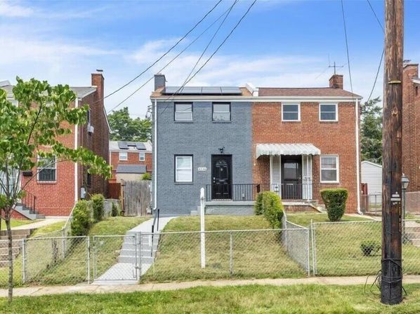 Front view of a two-story urban brick duplex—left unit painted gray with solar panels and black door, right unit red brick with a covered porch, shared chained-link fence, concrete steps, small lawns, sidewalk and a utility pole