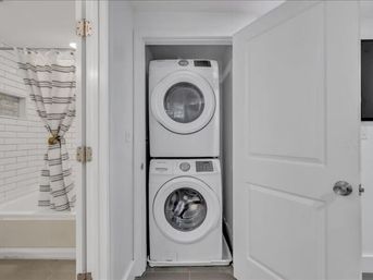 Stacked white front-load washer and dryer tucked into a narrow laundry closet beside a bathroom with white subway tile and a striped shower curtain.