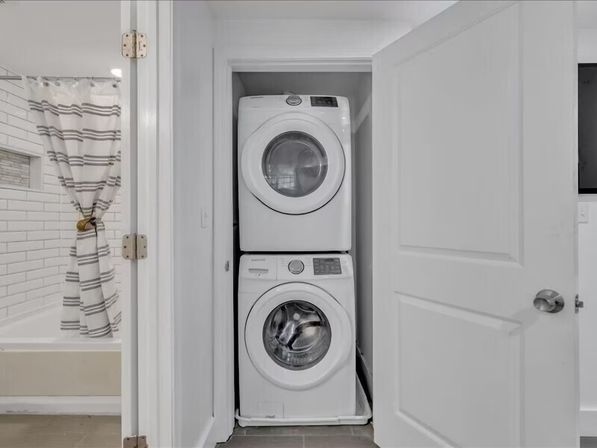 Stacked white front-load washer and dryer tucked into a narrow laundry closet beside a bathroom with white subway tile and a striped shower curtain.