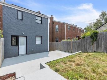 Sunny urban backyard with a gray-painted two-story brick house, white entry door and windows opening onto a concrete patio, small patchy lawn and a wooden privacy fence with neighboring red brick rowhouses visible
