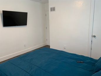Cozy minimalist bedroom with crisp white walls, wall-mounted flat-screen TV, dark hardwood floor and a blue comforter on the bed with a remote control