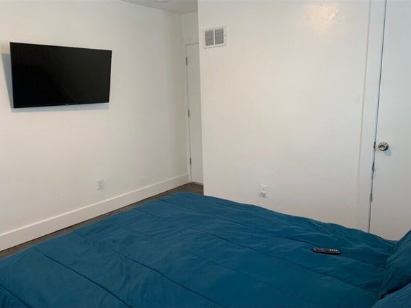 Cozy minimalist bedroom with crisp white walls, wall-mounted flat-screen TV, dark hardwood floor and a blue comforter on the bed with a remote control