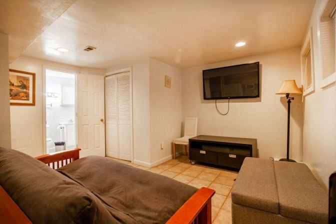 Cozy finished basement living area with futon, gray ottoman, wall-mounted TV above a media console, floor lamp, closet and open bathroom doorway on tiled floor.