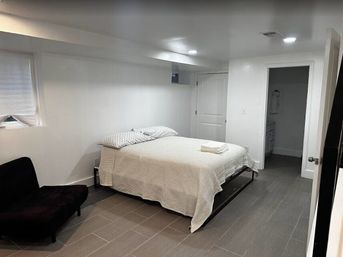 Cozy modern basement bedroom with a queen bed in white linens and patterned pillows, folded towels on the bed, recessed ceiling lights, small egress window, gray tile floor and a black accent chair