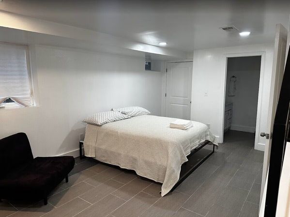 Cozy modern basement bedroom with a queen bed in white linens and patterned pillows, folded towels on the bed, recessed ceiling lights, small egress window, gray tile floor and a black accent chair