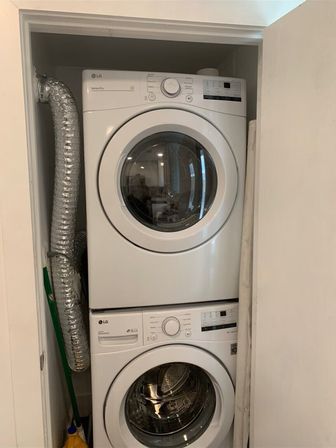 Stacked white front-load washer and dryer in a narrow laundry closet with a silver flexible vent hose, mop handle and yellow detergent bottle visible