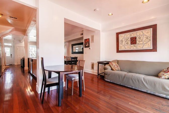 Open-plan living and dining area with glossy hardwood floors, round wooden table and chairs, neutral sofa with patterned pillows, framed wall art and recessed lighting.
