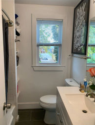 Compact white residential bathroom with vanity sink, toilet, shower curtain, and a window with blinds showing a leafy street view, framed black-and-white wall art and an orange rose on the countertop.