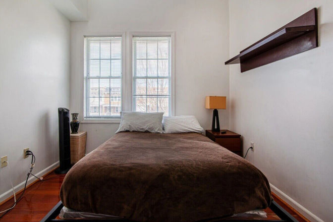Cozy urban apartment bedroom with a double bed in a brown duvet, two tall white-framed windows, hardwood floors, bedside table with lamp, wall shelf and a small tower fan