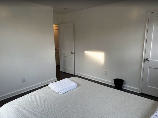 Minimal apartment bedroom with white walls and bedding, folded towels on the bed, two paneled doors, dark hardwood floor, small black waste bin and a rectangle of sunlight on the wall.