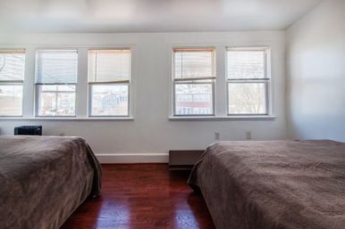 Sunlit city apartment bedroom with two twin beds in brown covers, dark hardwood floor, and a row of five windows with blinds overlooking neighborhood buildings.