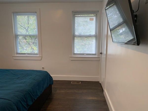 Minimal bedroom with teal duvet on a low bed, two windows with blinds letting in daylight, wall-mounted flat-screen TV, white walls and dark hardwood floor.