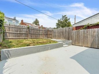 Compact urban fenced backyard with gray concrete patio, low painted cinderblock retaining wall and grassy raised bed, wooden privacy fence and neighboring rooftops under a blue sky