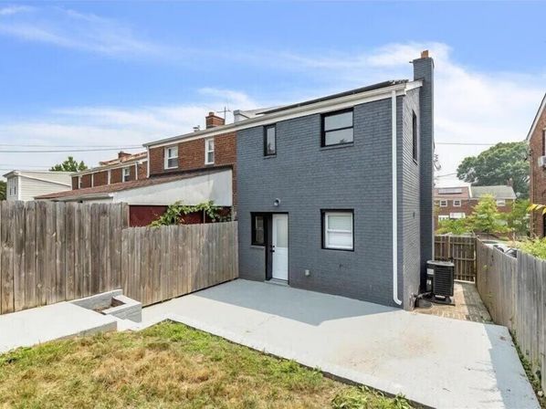 Rear view of a two-story gray brick rowhouse with a wooden privacy fence, small grassy yard, concrete patio, and outdoor AC unit under a clear blue sky.