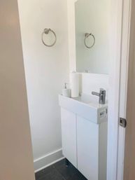 Small bright powder room with a compact white vanity sink, chrome faucet and towel ring, wall mirror, soap dispenser and paper towel roll — minimalist modern bathroom.